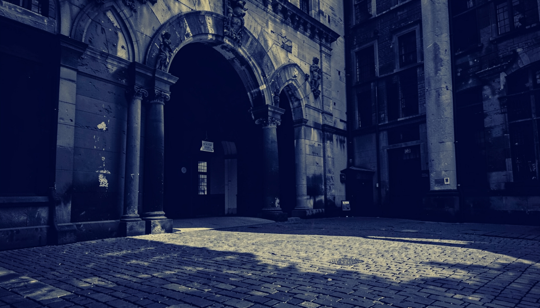 A duotone photograph in deep navy and muted gold tones showing the weathered stone archway and courtyard of a historic university building, with soft afternoon light casting long shadows across worn cobblestones
