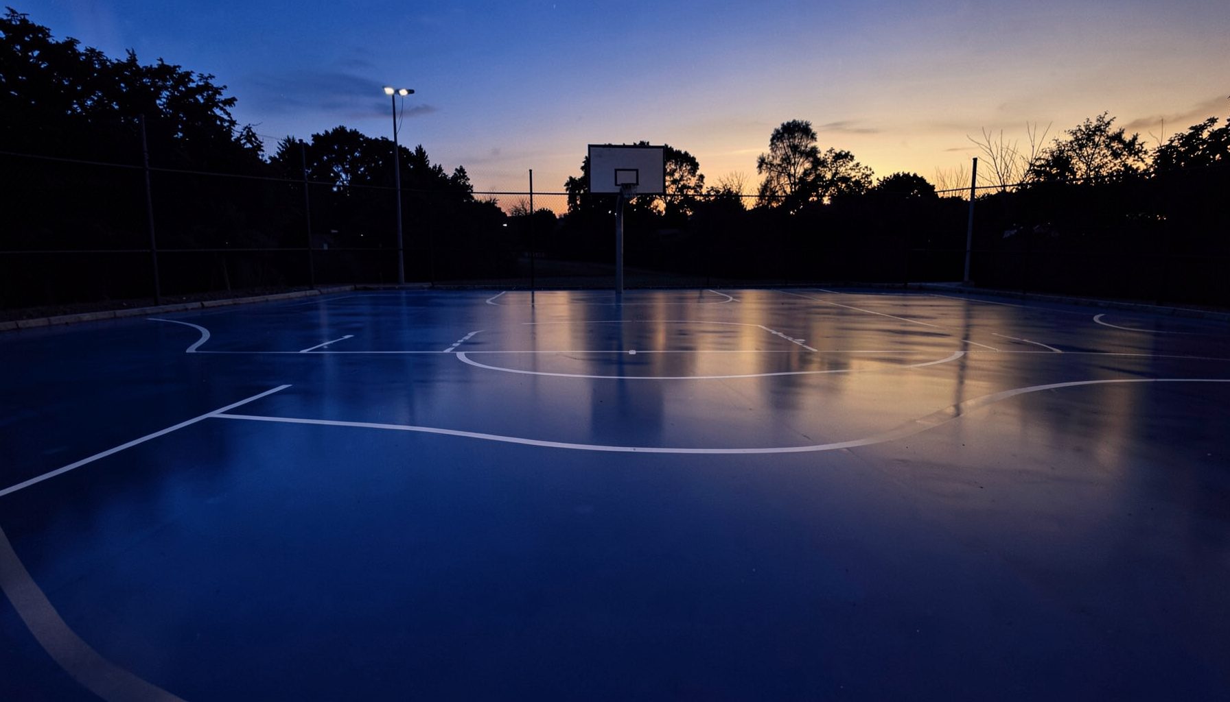 Wide duotone photograph of a freshly surfaced outdoor basketball court at dusk, deep blue and amber tones, smooth acrylic flooring reflecting soft light, empty court with crisp white boundary lines
