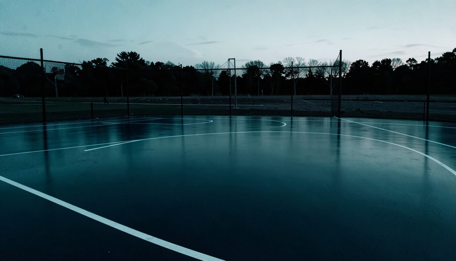 Wide duotone photograph of a freshly surfaced outdoor basketball court at dusk, deep navy and muted teal tones, smooth acrylic flooring reflecting soft ambient light, empty court with crisp white boundary lines stretching toward a distant hoop.